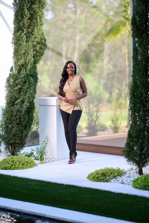 Full-length outdoor portrait in a tan blazer beside greenery and white columns