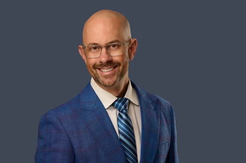 Professional headshot in a blue checked suit and striped tie against a gray backdrop
