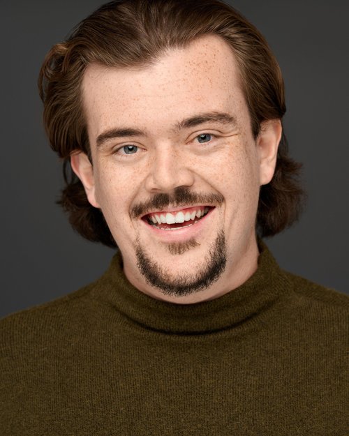Smiling talent headshot with a mustache and goatee in an olive turtleneck
